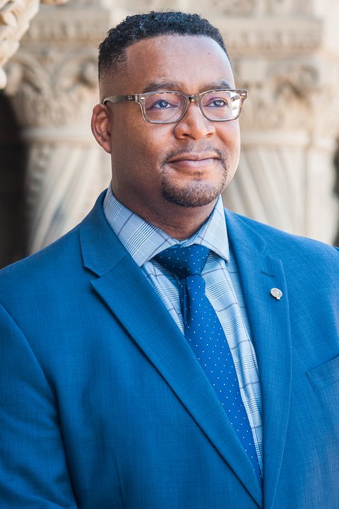 Kamal Hubbard, in a blue suit and glasses, in front of a carved stone doorway
