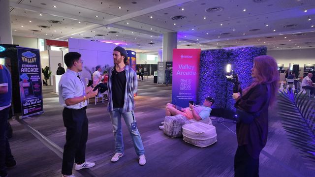 A camera operator with a light on her rig films two men talking beside the Valley Arcade banners, while a man lies on a beanbag behind them.