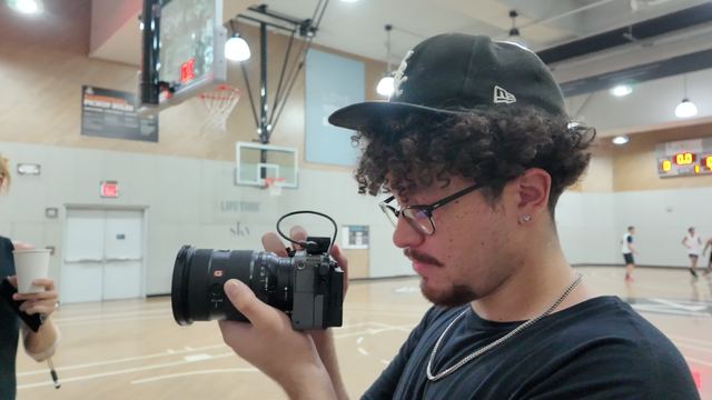 A photographer in a black cap and glasses looks down at his camera on an indoor basketball court, a hoop and players behind him.