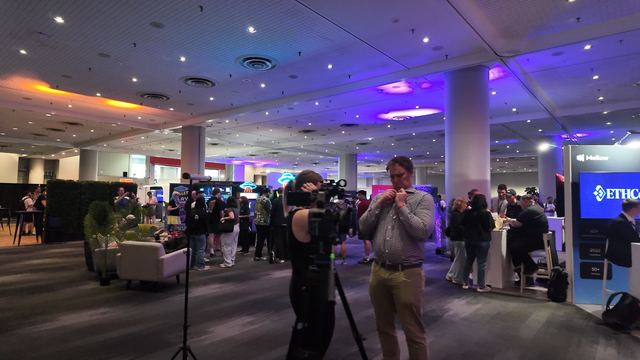 A camera operator at a video camera on a tripod lines up a man in a checked shirt clipping on a microphone, among the booths on the expo floor.