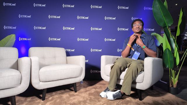A man with a conference badge clips on a microphone in an armchair on the ETHConf interview set, two empty chairs beside him and ETHConf logos on the wall behind.